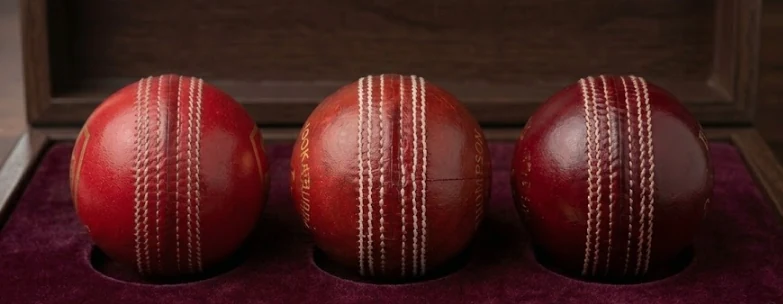 Three red Test cricket balls photographed seam-forward side by side — SG, Kookaburra and Dukes — each showing its six-row primary seam.